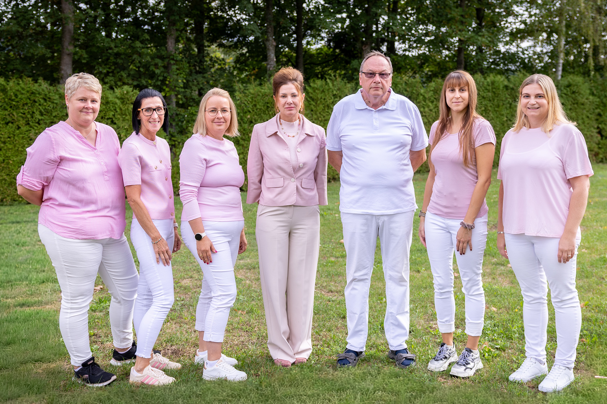 Gruppenfoto von sieben Personen im Freien vor einer grünen Hecke und Bäumen. Sechs Frauen und ein Mann stehen nebeneinander und blicken in die Kamera. Die Gruppe trägt farblich abgestimmte Kleidung in Rosa-, Weiß- und hellen Beigetönen.